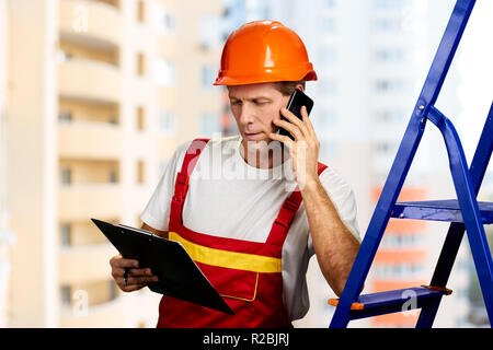 Ingegnere in elmetto parlando al telefono. Caposquadra parlando al telefono cellulare mentre guardando nella cartella nero, sfondo sfocato. Foto Stock