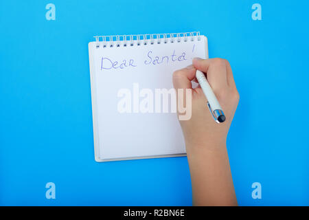 Lettera a Babbo Natale nel Blocco note con spazio per l'elenco. i bambini la mano a scrivere una lettera a Santa su sfondo blu vista superiore Foto Stock