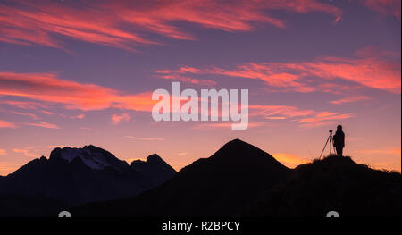 Silhouette di un fotografo sul picco di montagna contro la roccia e cielo viola con le Nuvole rosa al tramonto. Paesaggio con uomo, montagna cresta e colo Foto Stock