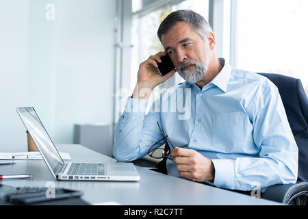 Executive senior imprenditore utilizzando il suo telefono mobile e parlando con qualcuno durante il funzionamento portatile in ufficio Foto Stock