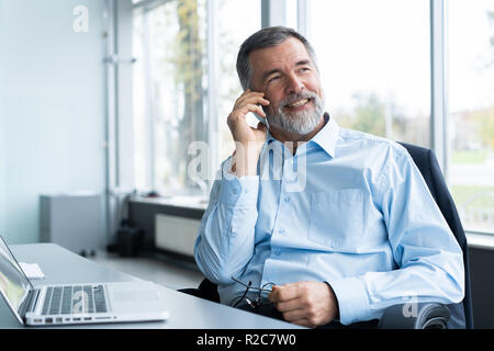 Executive senior imprenditore utilizzando il suo telefono mobile e parlando con qualcuno durante il funzionamento portatile in ufficio Foto Stock