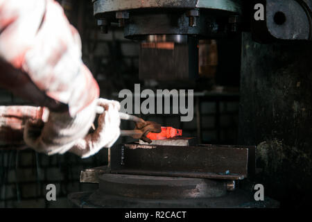 Fabbro lavora con red hot pezzo metallico della nuova ax sull'incudine e martello elettro presso la forge. Focus sull'incudine Foto Stock