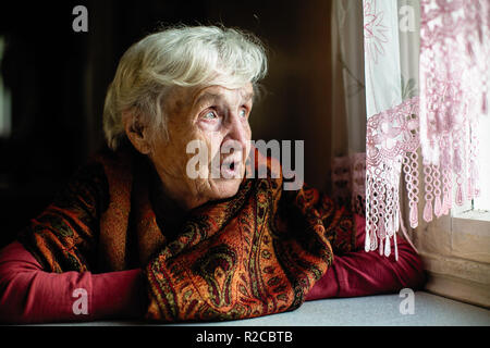 Donna anziana seduto in casa longingly guarda fuori dalla finestra. Foto Stock