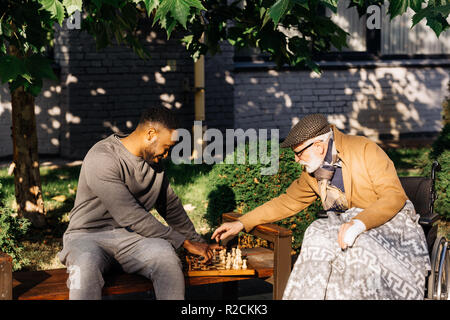 Senior uomo disabili in sedia a rotelle e african american nurse giocando a scacchi insieme sulla strada Foto Stock