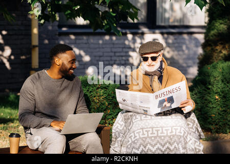 Felice senior uomo disabili business di lettura quotidiano in carrozzina mentre african american uomo utilizzando portatile su strada Foto Stock