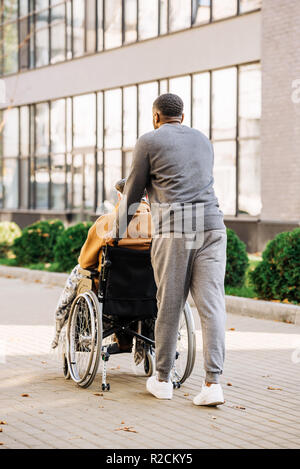 Vista posteriore del senior uomo disabili in carrozzella con plaid e cuidador riding da street Foto Stock