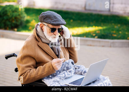 Bello senior uomo disabili in carrozzina tramite computer portatile con auricolari su strada Foto Stock