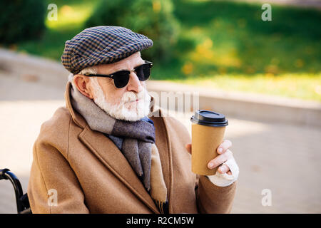 Close-up ritratto di senior uomo disabili in carrozzella tenendo la carta di tazza di caffè sulla strada Foto Stock