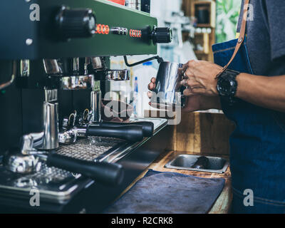 Professional barista che indossa jeans grembiule di latte per la cottura a vapore con acciaio inossidabile mug sulla macchina del caffè sul tavolo di legno al cafe'. Foto Stock