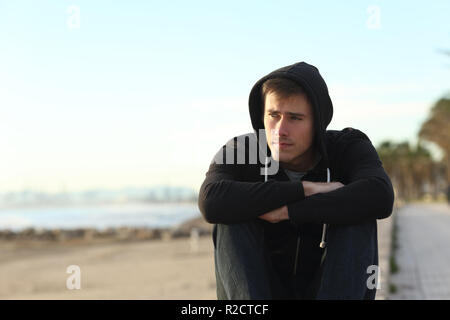 Vista anteriore verticale di un grave ragazzo adolescente che guarda lontano seduto sulla spiaggia Foto Stock