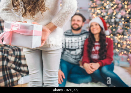 Bella foto della ragazza stnds nella parte anteriore dei genitori. Ella è presente. La ragazza si nasconde dietro di se stessa. I genitori seduti sul pavimento e guardare a lui. Hanno sorriso. Foto Stock