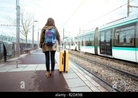 Ragazza turistico con uno zaino e una grande valigia gialla si erge sulla piattaforma e attende il treno Foto Stock