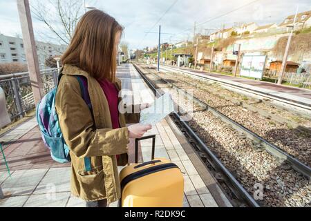 Ragazza turistico con uno zaino e una grande valigia gialla si erge sulla piattaforma e attende il treno Foto Stock