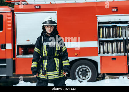 Firefighter femmina in uniforme di protezione permanente sulla strada con red fire carrello dietro Foto Stock