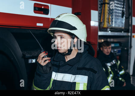 Donna pompiere parlando su radio portatile impostato con il collega dietro alla stazione dei vigili del fuoco Foto Stock