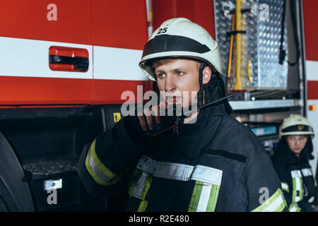 Messa a fuoco selettiva del vigile del fuoco in uniforme di parlare su radio portatile impostato con il collega dietro alla stazione dei vigili del fuoco Foto Stock