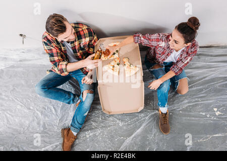 Vista aerea della coppia giovane tintinnio di bottiglie di birra e mangiare la pizza seduti sul pavimento in un nuovo appartamento Foto Stock