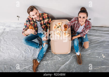 Vista aerea di felice coppia giovane con birra e pizza seduti sul pavimento e sorridente alla fotocamera nel nuovo appartamento Foto Stock