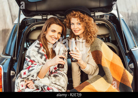 Messa a fuoco selettiva di felice Donne avvolte in coperte di contenimento delle bottiglie di soda seduta nel baule auto in strada urbana Foto Stock