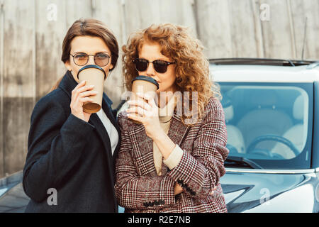 Ritratto di donna elegante amici in cappotti e occhiali da sole di bere il caffè nei pressi di auto in strada urbana Foto Stock