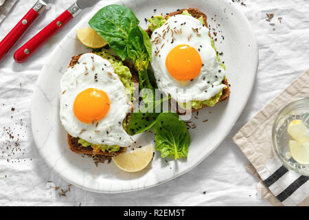 Vista in pianta sana avocado toast per la prima colazione o il pranzo con pane di segale, purea di avocado toast e uova fritte su sfondo bianco con un bicchiere di acqua di limone Foto Stock