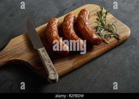 Tre deliziose salsicce con un rametto di rosmarino su una tavola di legno e il coltello su una tavola nera Foto Stock