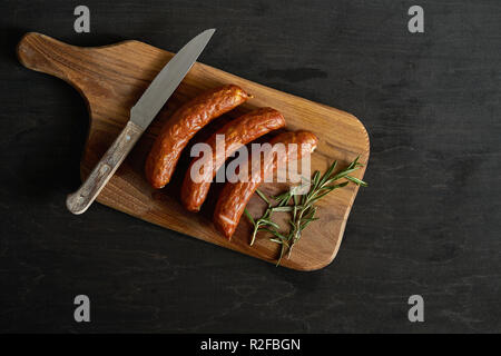 Vista superiore di tre deliziose salsicce con un rametto di rosmarino su una tavola di legno e il coltello su una tavola nera Foto Stock