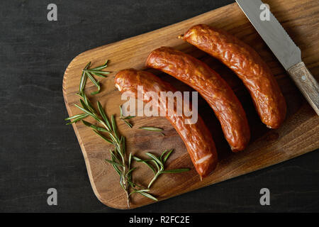 Vista superiore di tre deliziose salsicce con un rametto di rosmarino su una tavola di legno e il coltello su una tavola nera Foto Stock