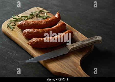 Tre deliziose salsicce con un rametto di rosmarino su una tavola di legno e il coltello su una tavola nera Foto Stock