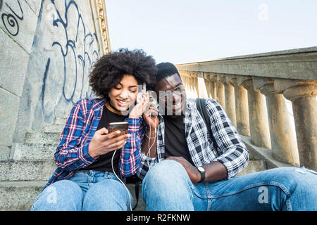 Allegro felice africann nero giovane seduto togethe in amicizia ascoltando la musica da un telefono moderno e condividendo le cuffie come partner. sorridente e divertirsi Foto Stock