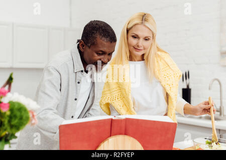 Felice interracial giovane con il libro di cucina la cena insieme in cucina Foto Stock