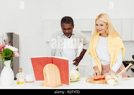 Felice interracial giovane cucinare la cena insieme e la lettura di ricetta nel ricettario di cucina Foto Stock