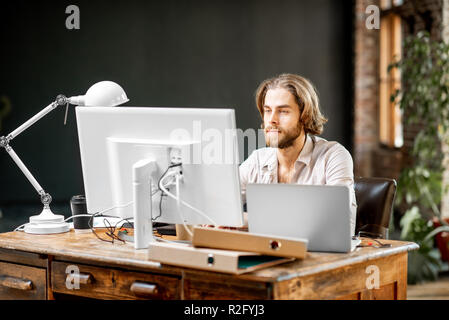 Uomo bello lavorare con laptop e computer seduti a tavola in casa moderna o un ufficio interno Foto Stock