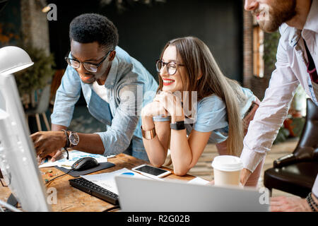 Un gruppo di giovani la multietnicità collaboratori vestito casualmente lavorando insieme focalizzata sul monitor del computer in ambienti interni Foto Stock