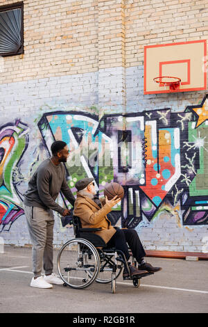 Senior uomo disabili in sedia a rotelle e felice african american uomo giocare a basket insieme sulla strada Foto Stock