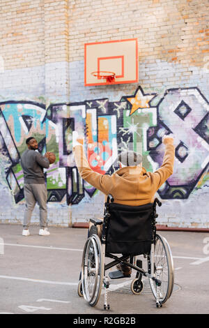 Vista posteriore di celebrare senior uomo disabili in carrozzella guardando african american uomo giocare a basket su strada Foto Stock