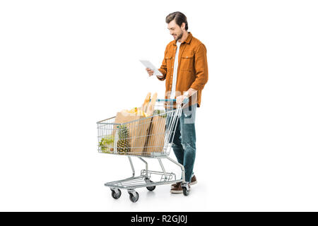 Uomo bello controllando la lista della spesa sulla tavoletta digitale e portante carrello isolato su bianco Foto Stock