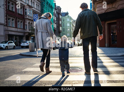 Una vista posteriore del piccolo bimbo ragazzo con i genitori attraversare una strada all'aperto in città. Foto Stock