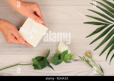 Donna di mani tenendo una confezione regalo su grigio Sfondo di legno con splendidi fiori e verde leavs. Concetto di dare un regalo in vacanza o compleanno. Parte superiore Foto Stock