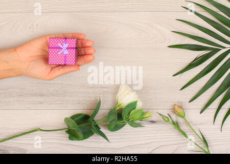 Donna di mani tenendo una confezione regalo su grigio Sfondo di legno con splendidi fiori e verde leavs. Concetto di dare un regalo in vacanza o compleanno. Parte superiore Foto Stock