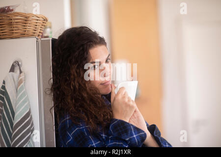 Donna appoggiata contro un frigorifero di bere una tazza di caffè Foto Stock