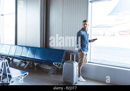 Uomo con argento Valigia controllo telefono e in attesa vicino alla finestra in aeroporto Foto Stock