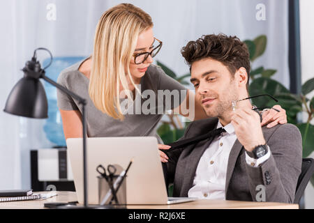 Ritratto di imprenditrice tirando i colleghi cravatta al lavoro con il computer portatile in ufficio, flirtare e ufficio concetto romantico Foto Stock
