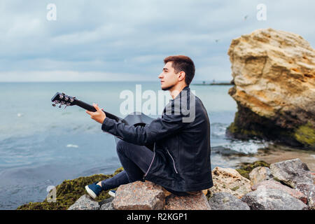 Giovane uomo con la chitarra acustica a giocare da solo sulla spiaggia circondata da rocce sul giorno di pioggia Foto Stock