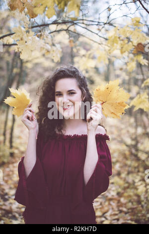 Bellissima ragazza con ricci capelli scuri in un marron top in un autunno park, sorridente verso la telecamera e tenere foglie di acero Foto Stock