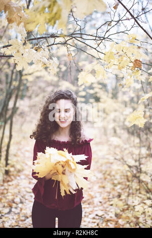 Bellissima ragazza con ricci capelli scuri in un marron top in un autunno park holding foglie di acero e sorridente Foto Stock