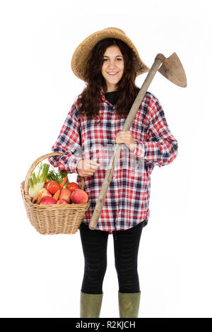 Giovane agricoltore con un braccio che tiene una cesta di frutta e verdura, con gli altri una zappa, indossa un cappello di paglia, controllati e maglietta verde in gomma Foto Stock