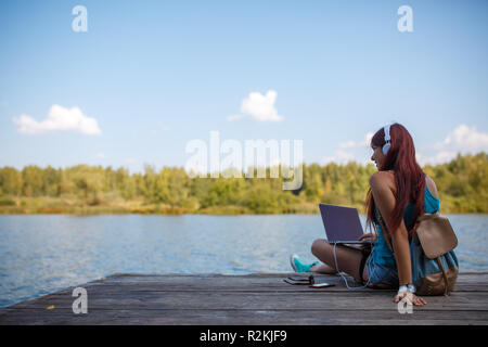 Foto sul retro della giovane bruna nelle cuffie con zaino e laptop in mani seduto sulla riva del fiume al pomeriggio estivo Foto Stock