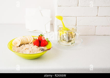 Farina di avena porridge con banana e fragola in verde ciotola con tè caldo e sana colazione su sfondo chiaro con spazio di copia Foto Stock