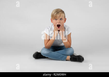 Piccolo ragazzo biondo si siede con le gambe incrociate sul pavimento ed è sorpreso con la bocca aperta contro uno sfondo bianco Foto Stock